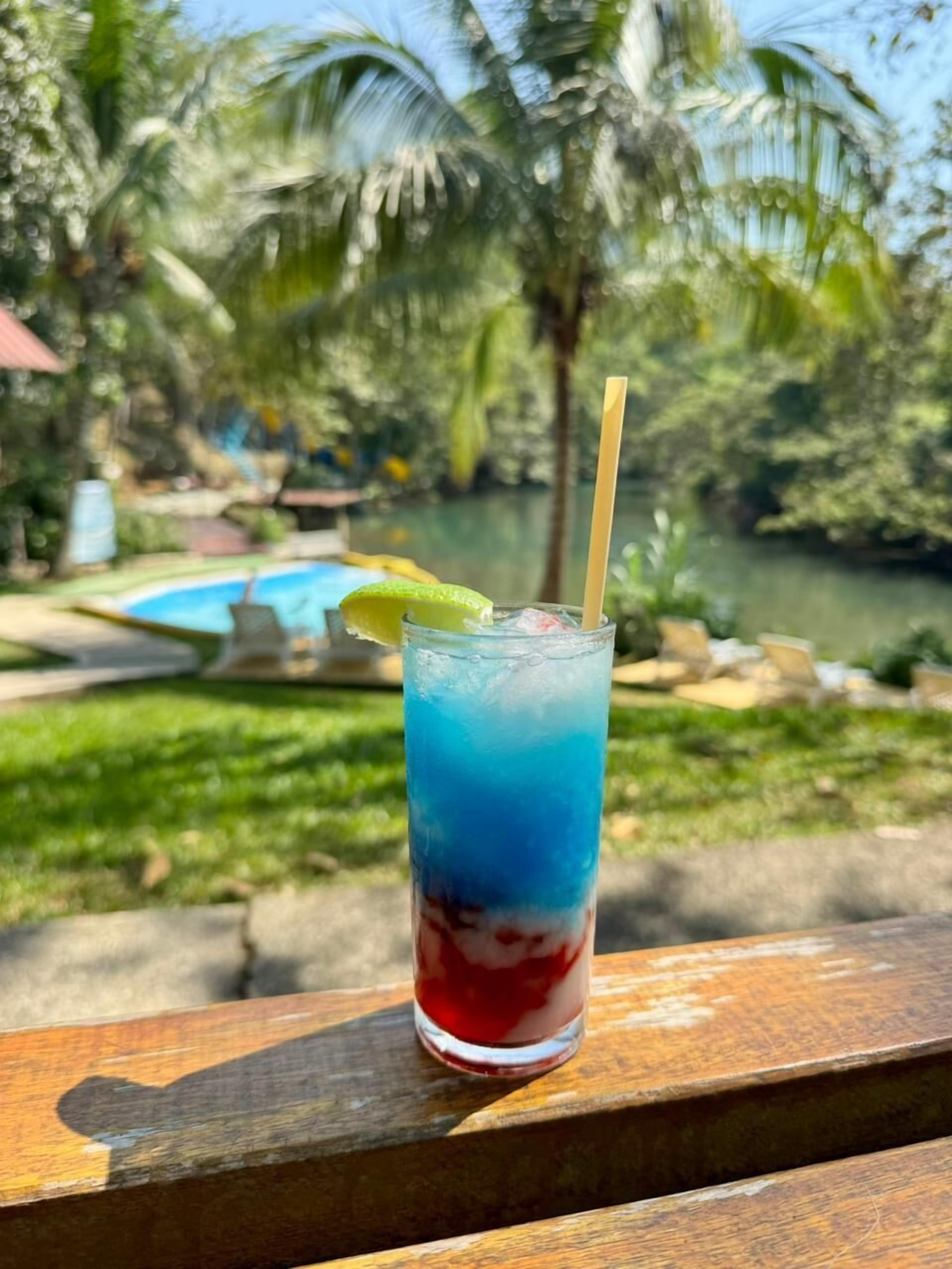A tropical drink with a straw, a lime wedge, and a glass of blue and red liquid, placed on a wooden surface with a pool and palm trees in the background.