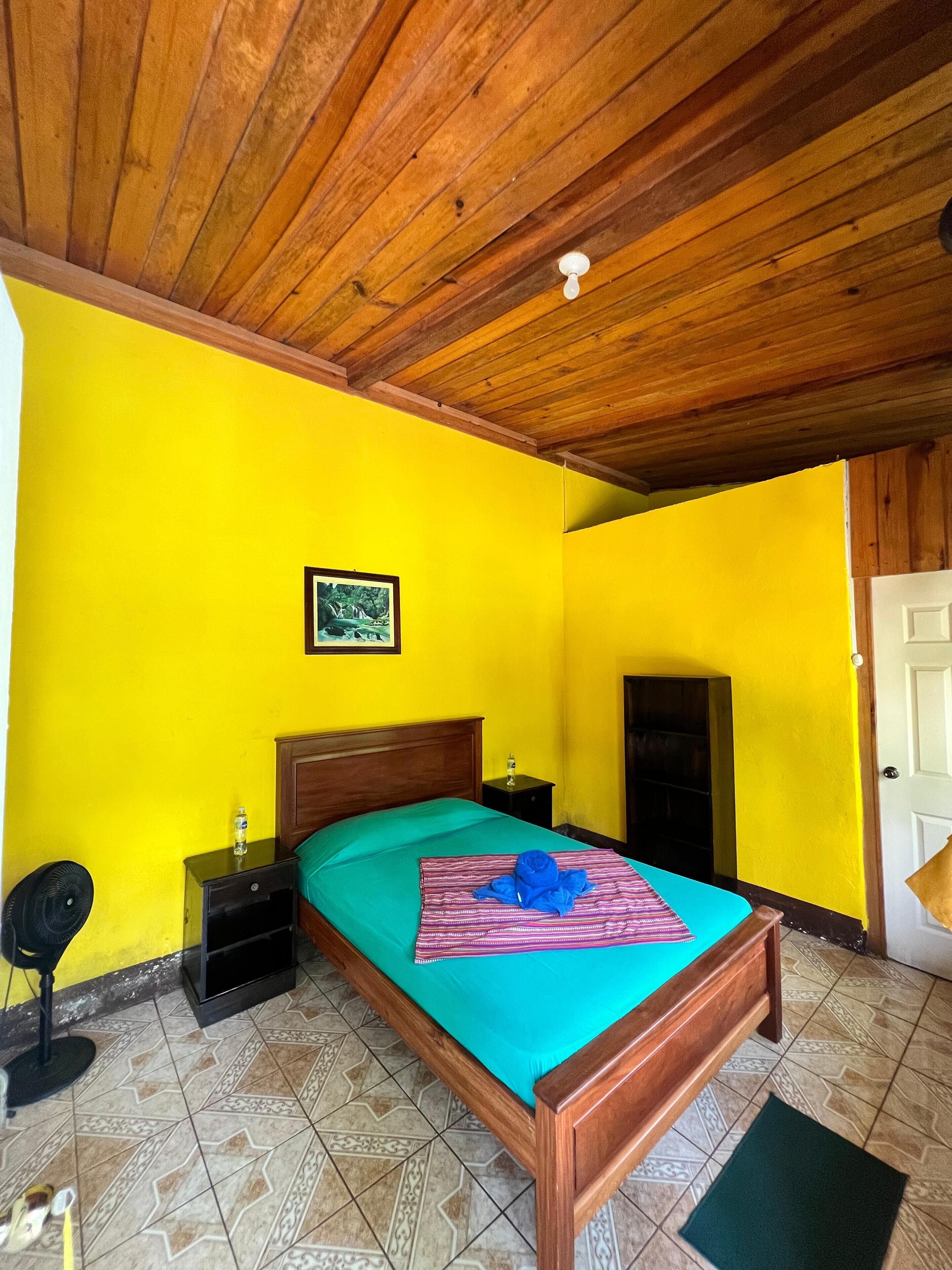 A bedroom with a wooden ceiling, a bed with a green bedspread, a framed picture on the wall, and a nightstand with a lamp.