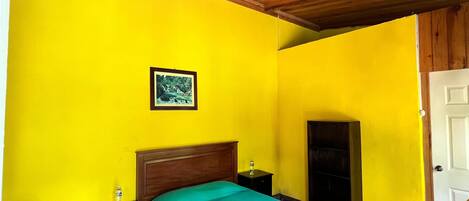 A bedroom with a wooden ceiling, a bed with a green bedspread, a framed picture on the wall, and a nightstand with a lamp.
