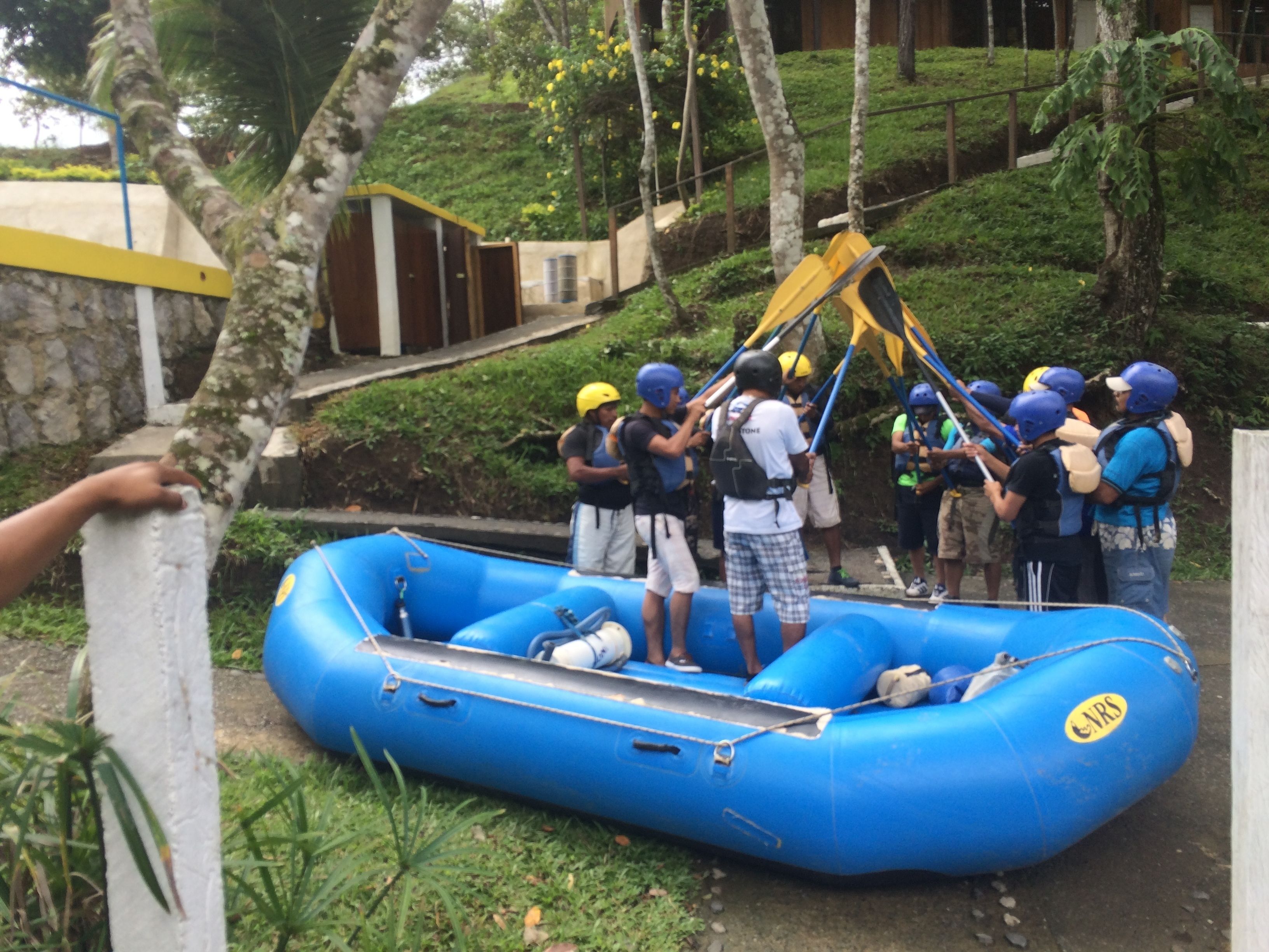 A group of people in a blue inflatable raft, some wearing helmets, near a building with a yellow door.
