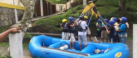 A group of people in a blue inflatable raft, some wearing helmets, near a building with a yellow door.