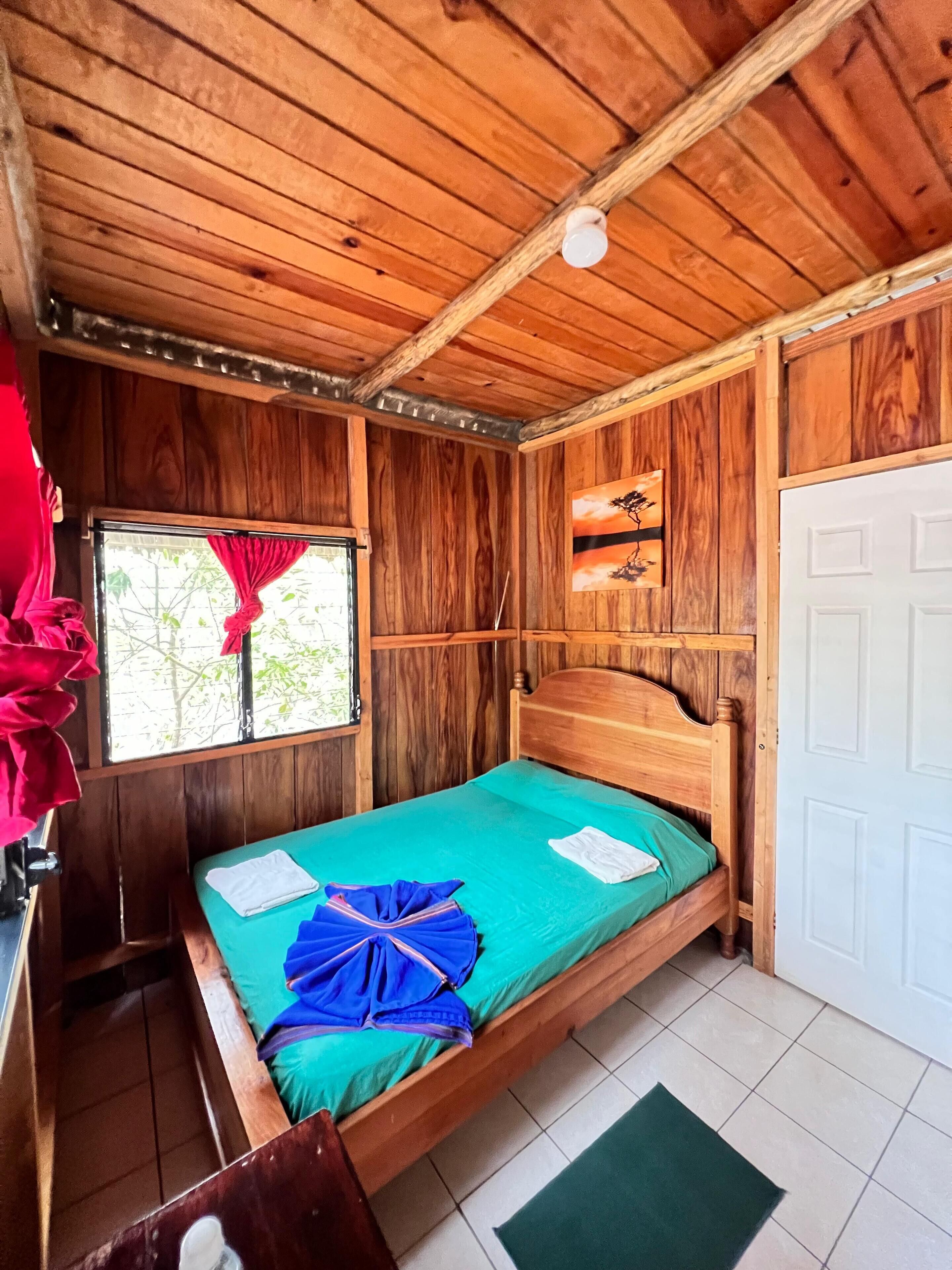 A wooden room with a bed, a window with red curtains, a painting, and a white door.