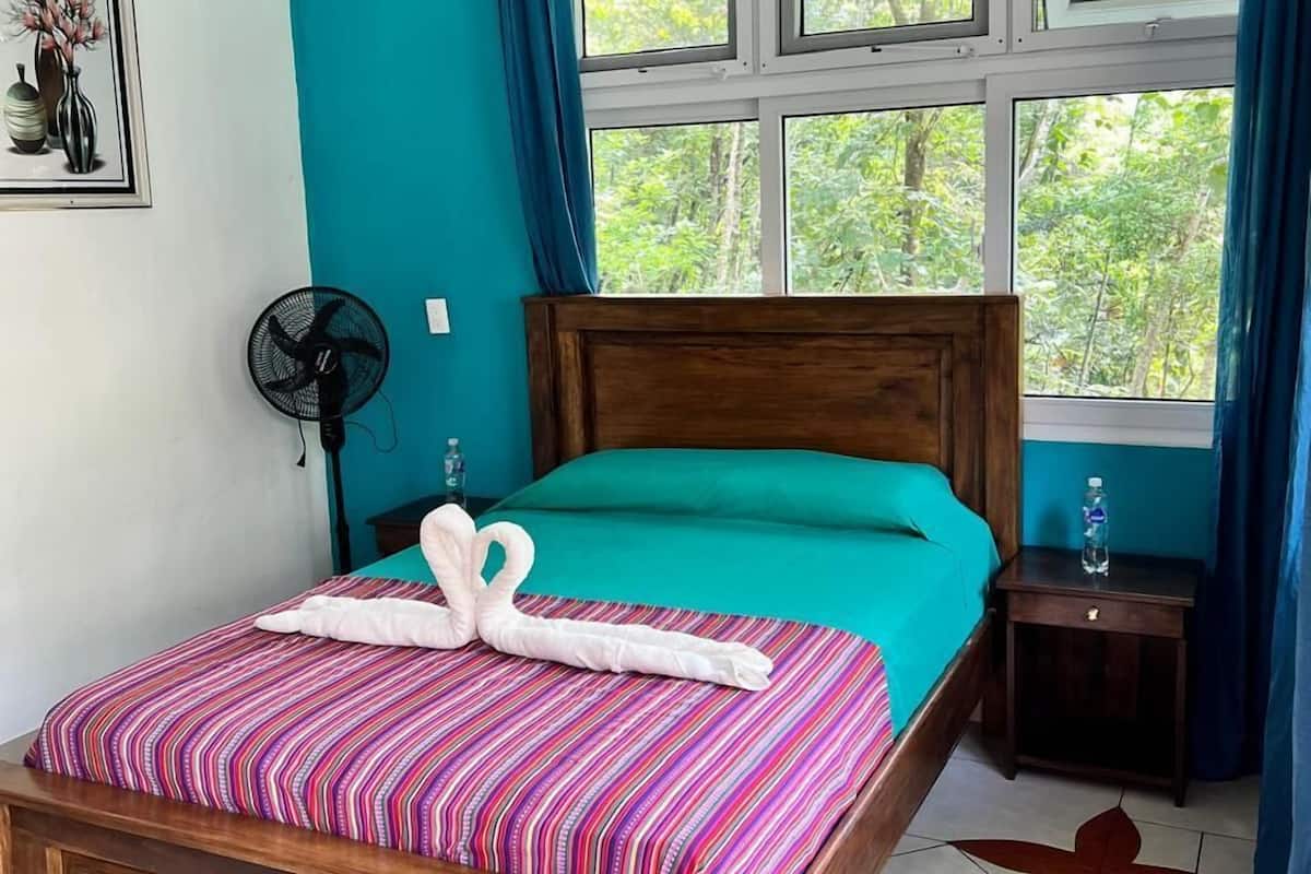 Une chambre avec un lit en bois, des murs de couleur turquoise, une fenêtre avec des rideaux, une table de chevet avec une bouteille d’eau et un ventilateur.