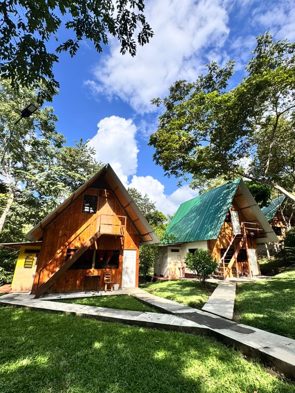 A rustic cabin with a sloped roof and a wooden exterior, surrounded by lush greenery and a clear blue sky.