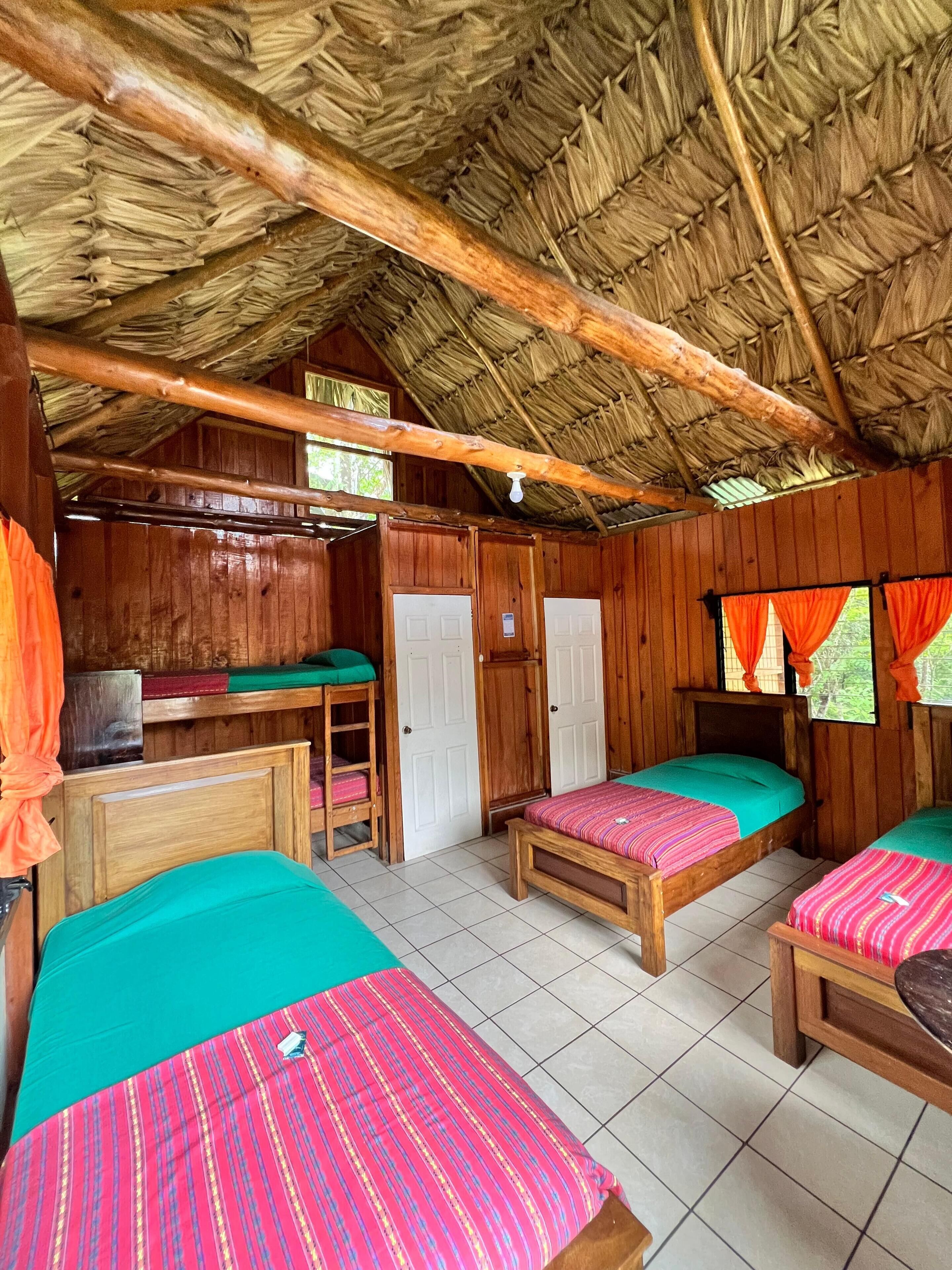 Habitación con cuatro camas, un escritorio y una silla. La habitación tiene techo de paja y paredes de madera.