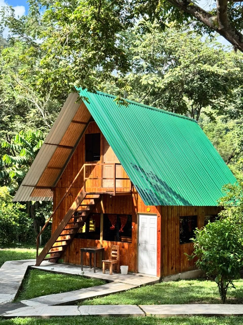 A wooden cabin with a green roof, a staircase leading to a deck, and a small outdoor seating area.