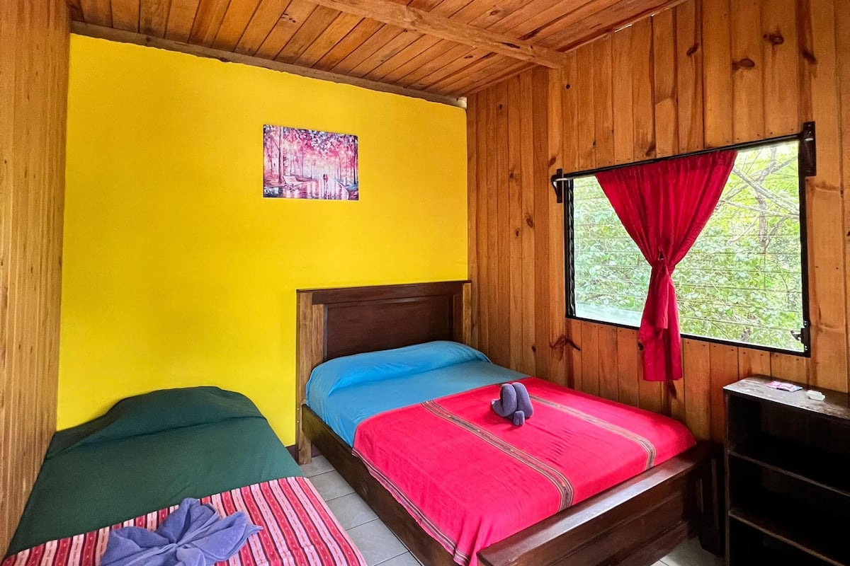 Une chambre avec deux lits, un ventilateur de plafond, une fenêtre avec des rideaux rouges et un plafond en bois.