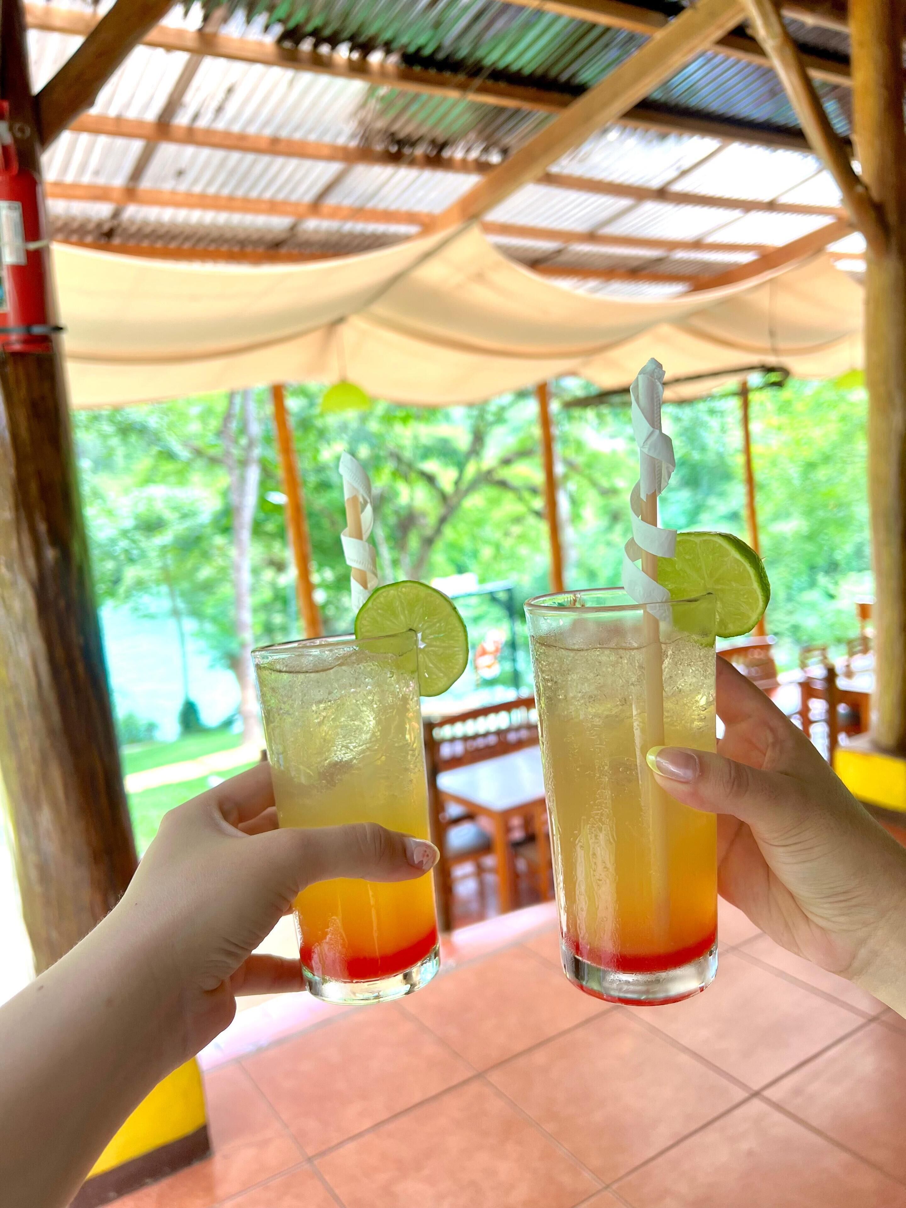 Two people holding glasses of a layered drink with a lime garnish. The setting appears to be an outdoor area with a thatched roof and a view of greenery in the background.