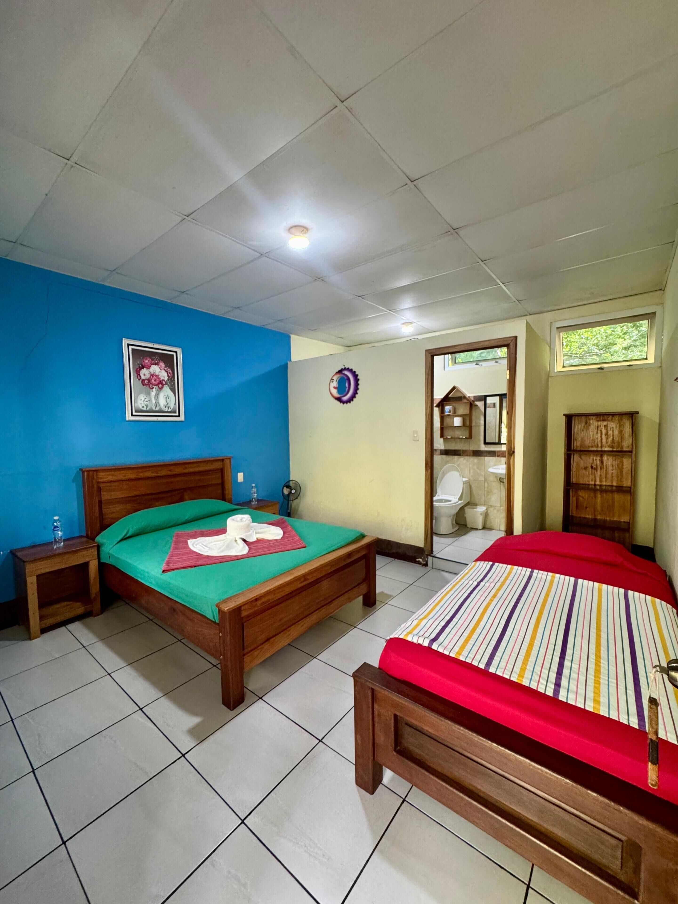A room with two wooden beds, a blue wall, a framed picture, a ceiling fan, and a window.
