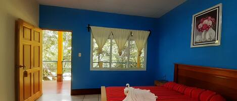 Un dormitorio con cama, ventana con cortinas, puerta y un cuadro en la pared.