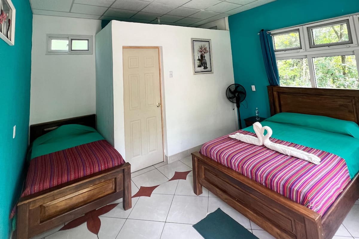 Une pièce avec deux lits en bois, un ventilateur de plafond, une fenêtre avec des rideaux et une porte.
