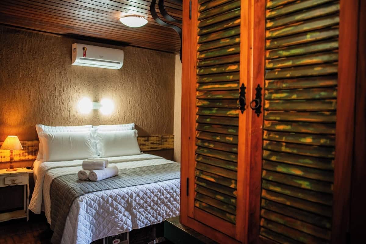 Um quarto com uma cama, uma mesa de cabeceira, um aparelho de ar condicionado e um guarda-roupa de madeira com portas venezianas.