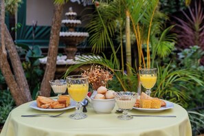 Mesa com itens de café da manhã, incluindo doces e suco de frutas, em ambiente externo com uma fonte e vegetação ao fundo.