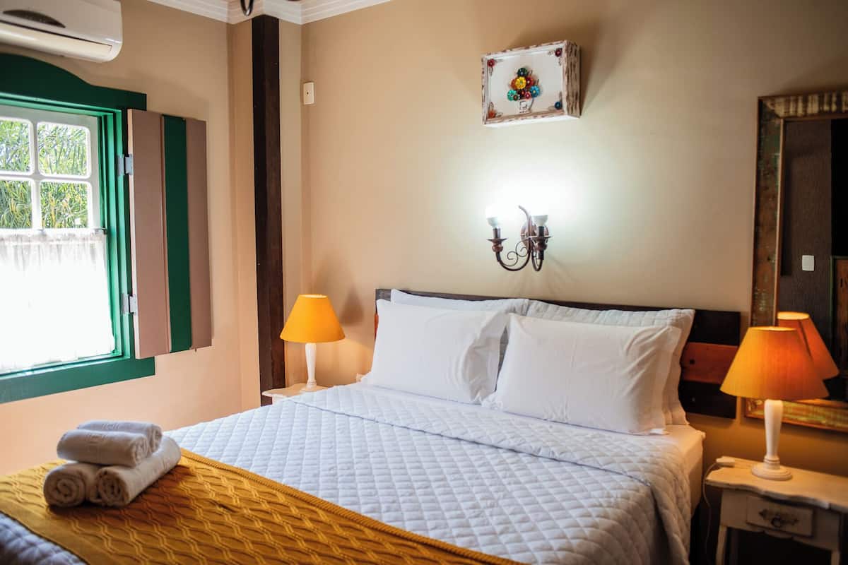 Cama bem arrumada com lençóis brancos, duas luminárias de cabeceira, luminária na parede e janela com persianas verdes.
