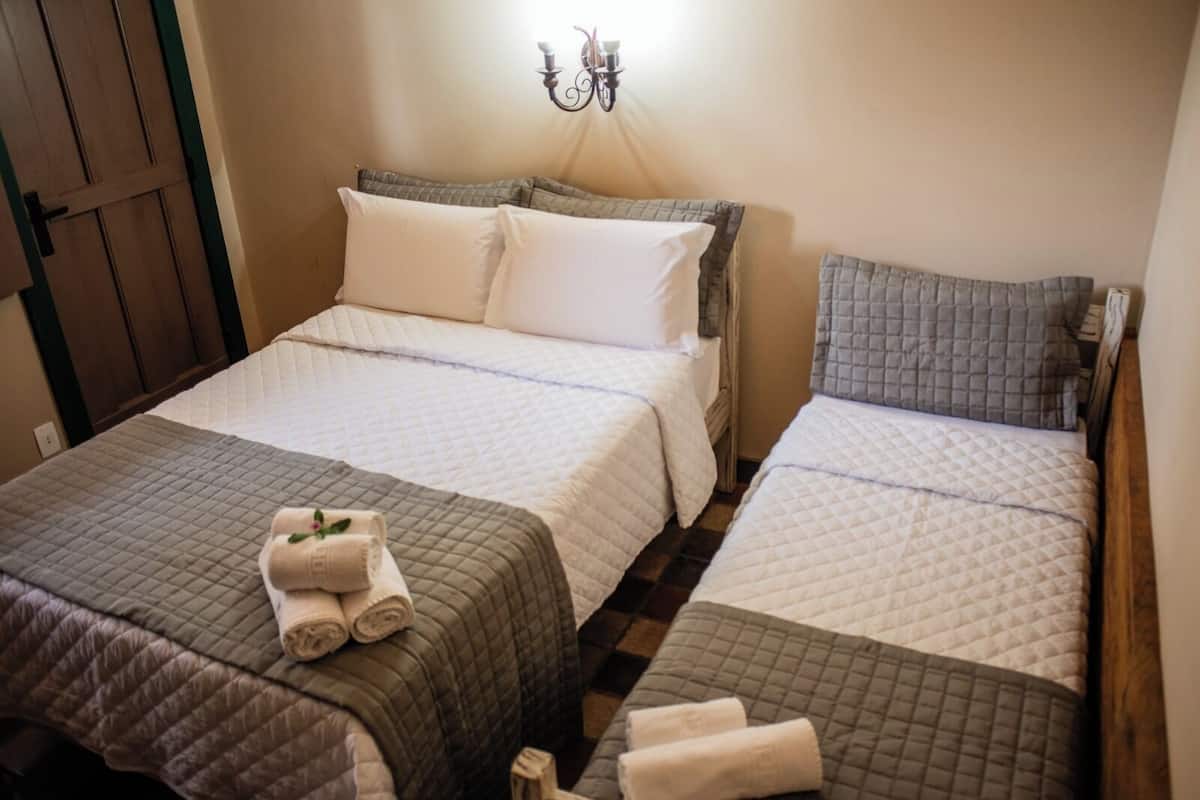 Quarto de hotel com duas camas, cada uma com colcha acolchoada e travesseiros. Há um candeeiro fixado na parede sobre as camas.