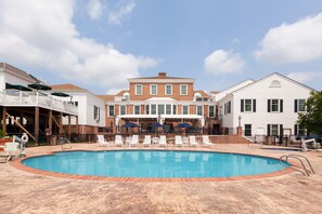 A swimming pool with lounge chairs and umbrellas, surrounded by brick buildings with white trim and a large central building with a gabled roof. - Virginia Crossings Hotel, Tapestry Collection by Hilton (Glen Allen)