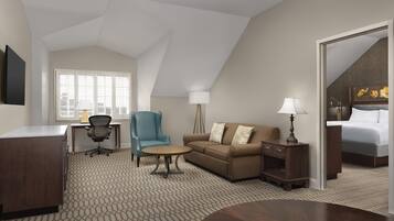A hotel room with a desk, a blue armchair, a brown sofa, a round coffee table, a bedside table with a lamp, and a bed with a floral painting.