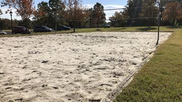 A sandy sports field with a volleyball net, surrounded by trees and parked cars.