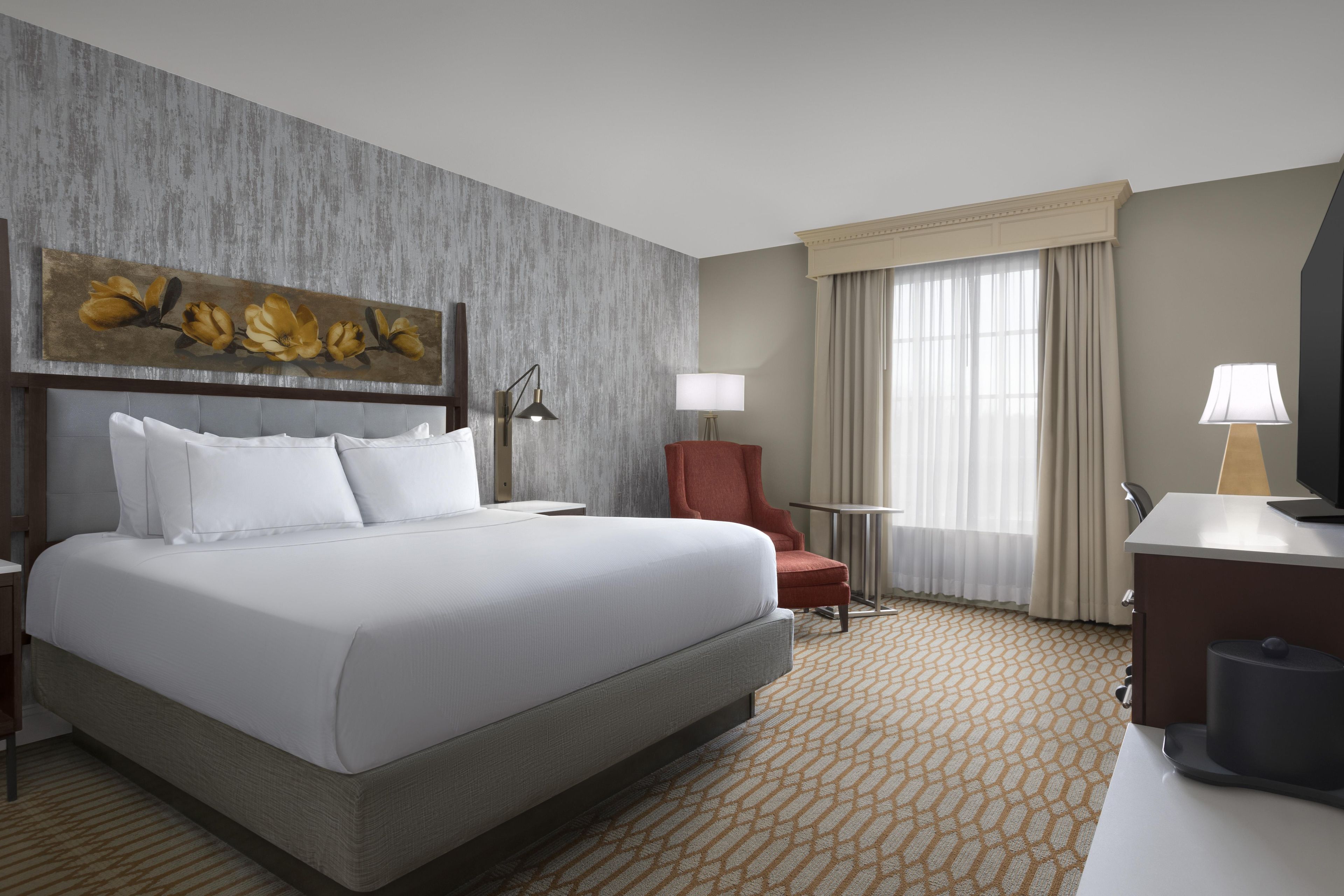 A hotel room with a large bed, a desk, a chair, a lamp, and a painting on the wall.