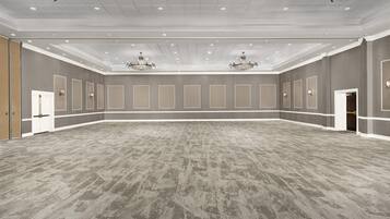 A spacious room with a grey carpet, white walls, and a ceiling with recessed lighting and chandeliers.