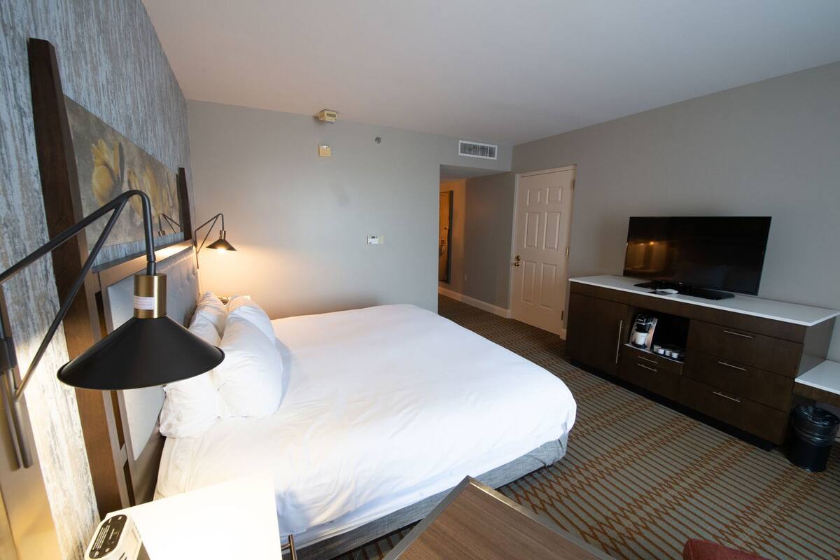 A hotel room with a bed, a TV, a desk with a lamp, a dresser, and a door leading to another room.