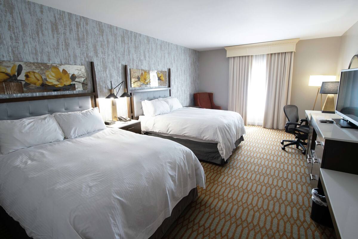 A hotel room with two beds, a desk with a computer, a chair, and artwork on the wall.