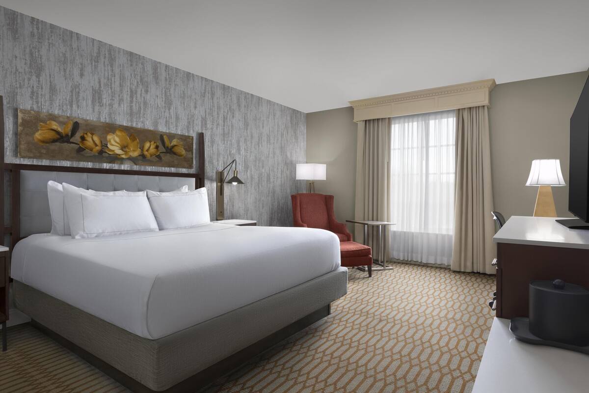 A hotel room with a large bed, a desk, a chair, a lamp, and a painting on the wall.