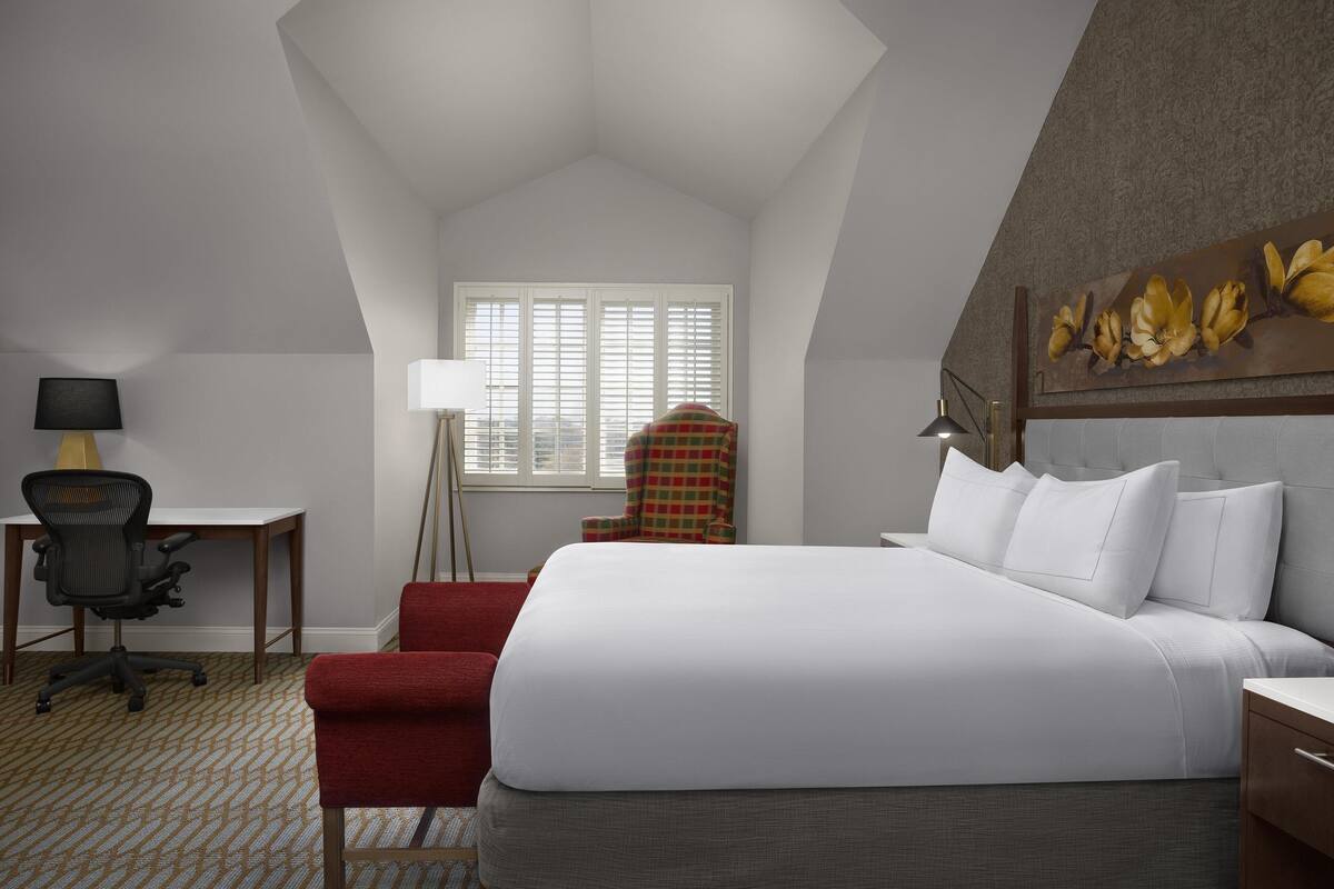 A hotel room with a large bed, a desk with a chair, a red armchair, and a floor lamp.