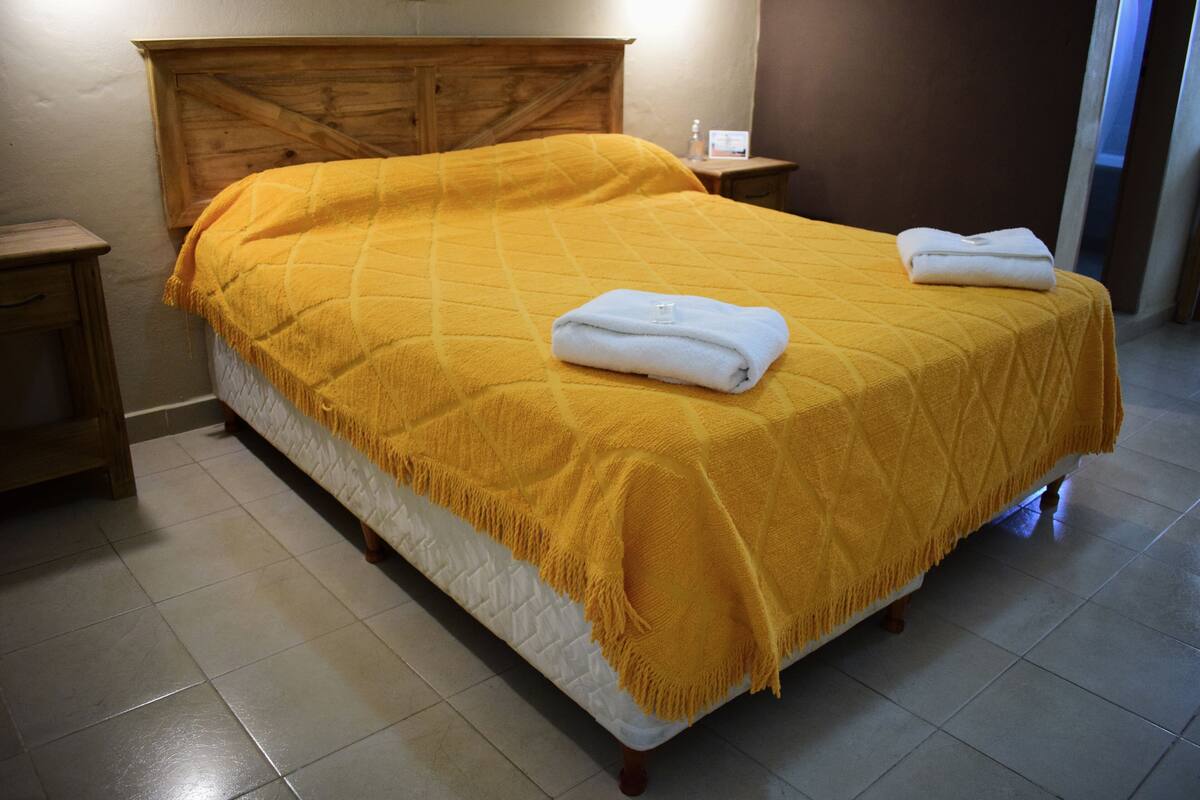 Cama com cobertor amarelo e duas toalhas dobradas, cabeceira de madeira, mesas de cabeceira e piso ladrilhado.
