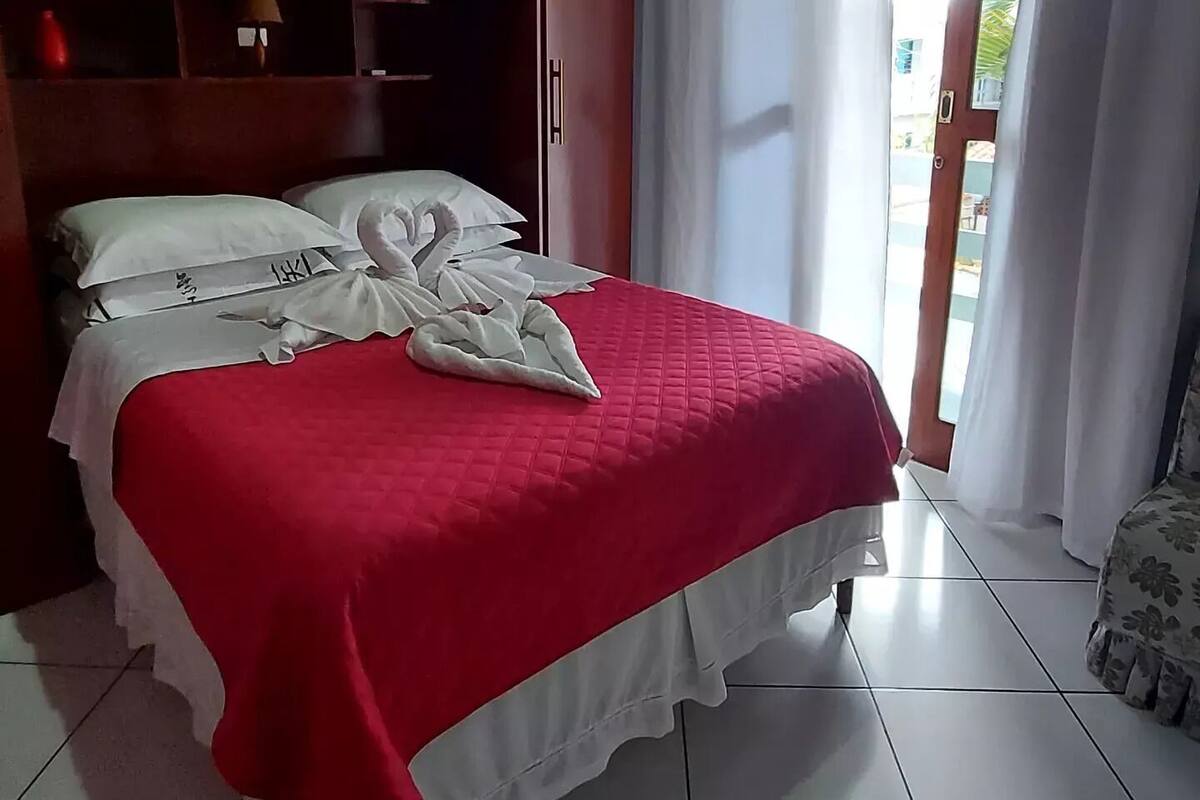 A hotel room with a bed, a wooden wardrobe, a small table, and a view of the outside through a glass door.