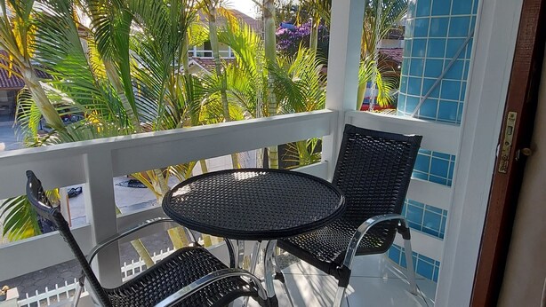 A covered outdoor seating area with two black metal chairs and a table, surrounded by a white railing, with a view of palm trees and a building in the background. - Pousada Estrela Guia (Florianópolis)
