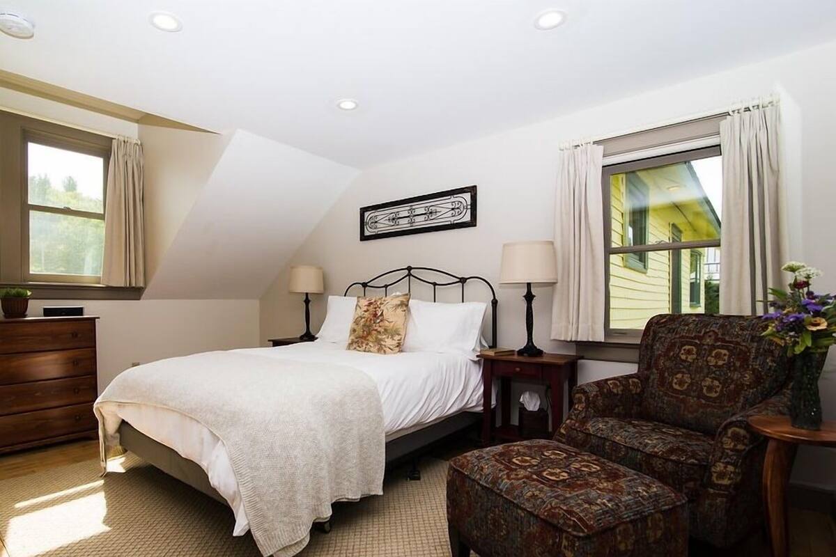 Un dormitorio con cama, dos mesitas de noche, una silla, cómoda y ventana con cortinas.