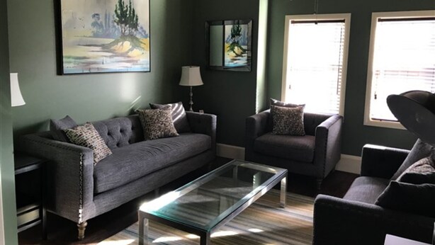 Ein Wohnzimmer mit Sofa, Sessel und Couchtisch. An der Wand hängen zwei Gemälde und eine Deckenleuchte.