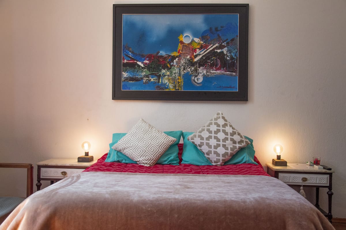 A neatly made bed with a red and white bedspread, two patterned pillows, and a framed abstract painting above it.