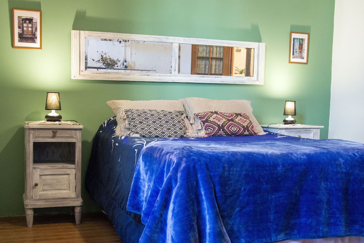 A bedroom with a bed, two bedside tables, a mirror, and a window with a view of a building.