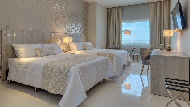 A hotel room with two beds, a desk, a chair, a TV, and a view of the city. - Hotel Dorado Plaza Alto Prado (Barranquilla)