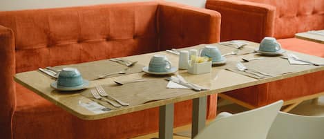 A dining area with a long table set for four, orange upholstered sofas, and white chairs.