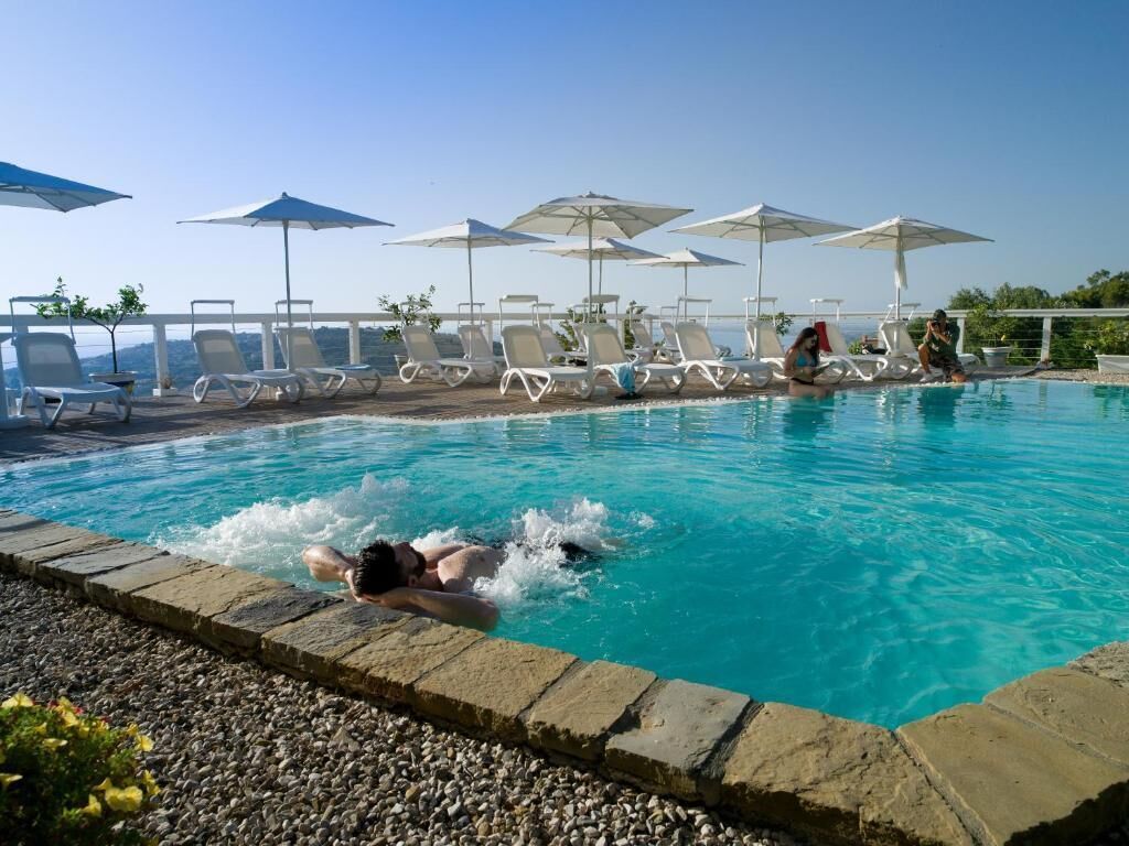 Una piscina con sillones de descanso y sombrillas, una persona nadando y un cielo despejado.