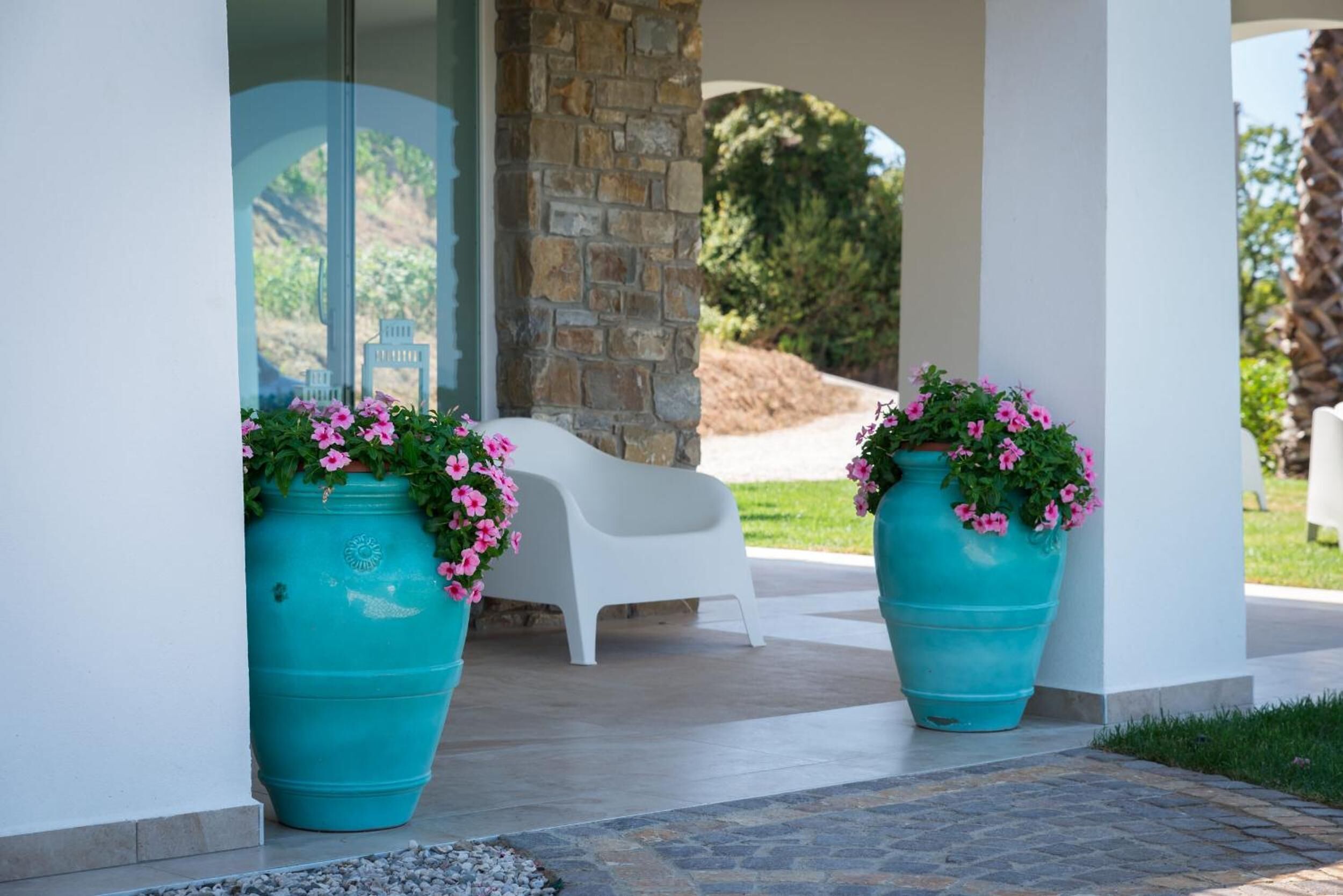 Un espacio exterior cubierto con dos macetas turquesas grandes con flores rosas, una silla de plástico blanco y un pilar de piedra.