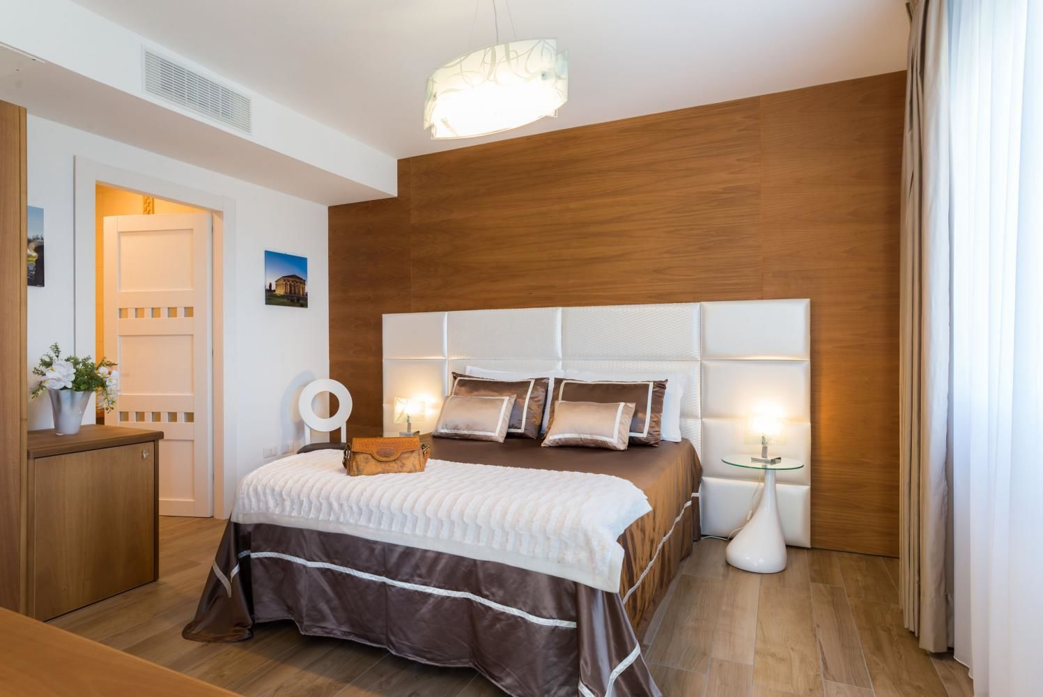 Quarto moderno com cabeceira de madeira, cama com roupa de cama branca e castanha, mesas de cabeceira com luminárias e chão de madeira.