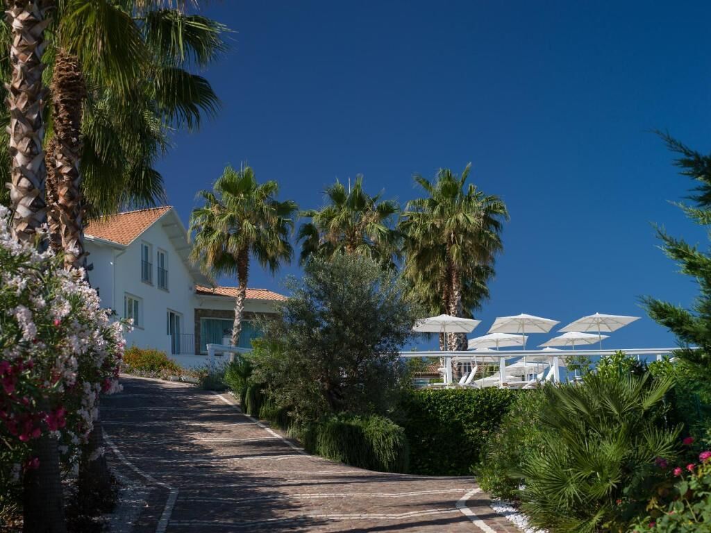 Un bâtiment blanc surmonté d’un toit recouvert de tuiles rouges, entouré de palmiers et bordé d’un chemin pavé menant à un espace extérieur abrité, aménagé avec des parasols.