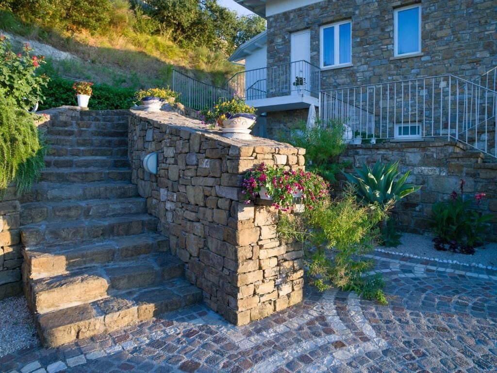 A stone staircase leading to a house with a balcony, surrounded by potted plants and flowers.