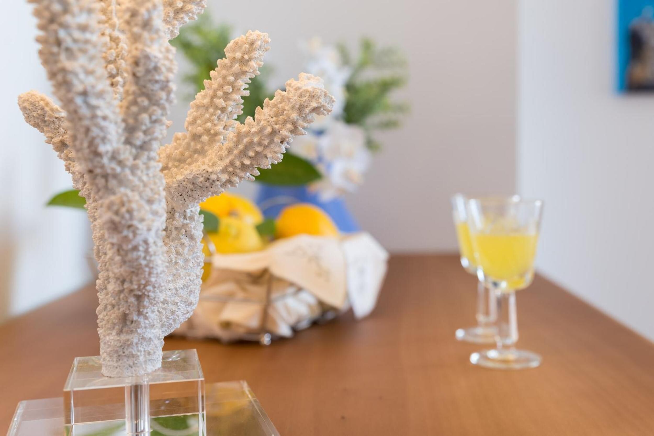 Eine transparente Skulptur aus Glas, die ein Korallenriff darstellt, auf einem klaren Sockel. Daneben ein Glas mit gelber Flüssigkeit und im Hintergrund ein Korb mit Früchten und Blumen.