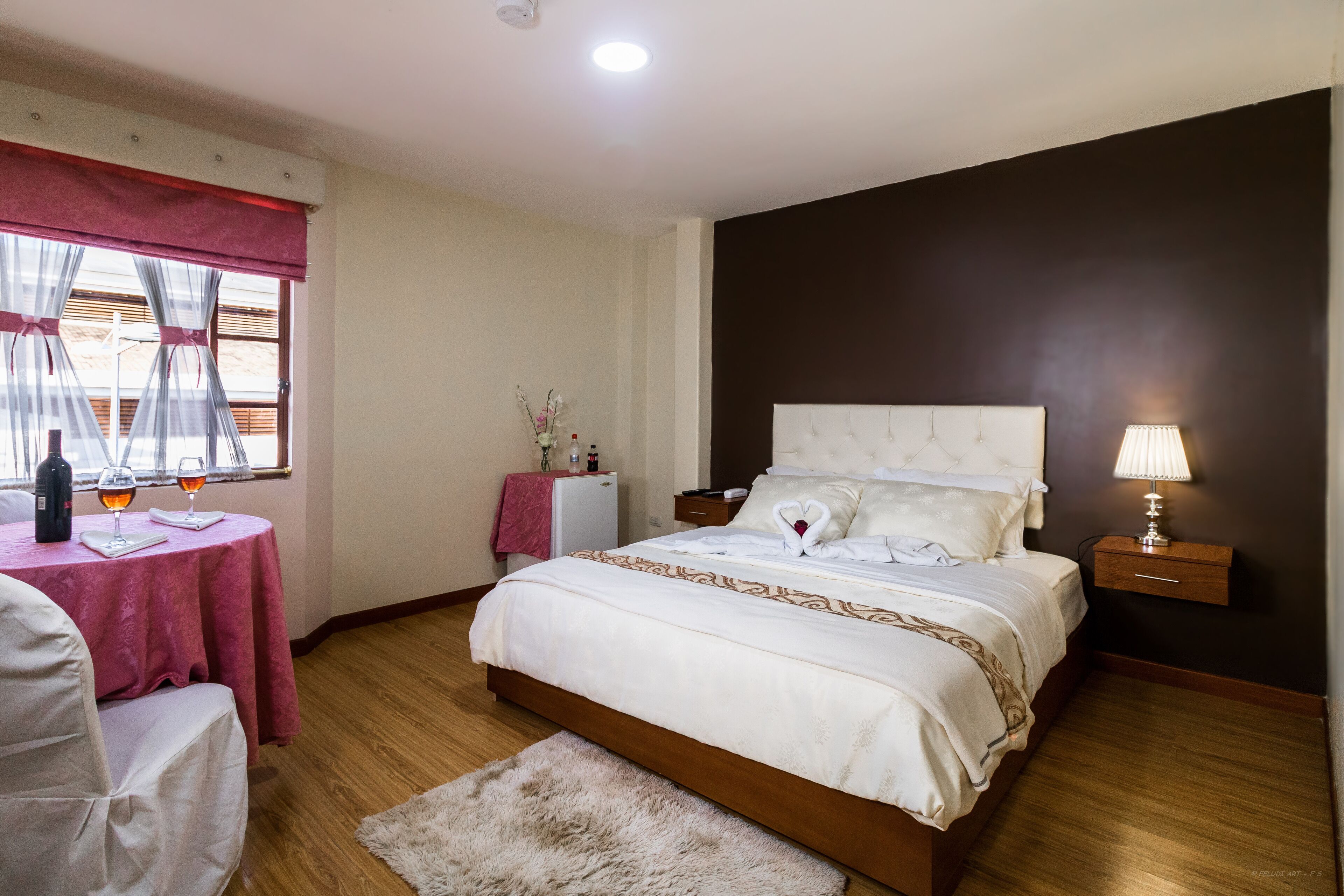 A bedroom with a large bed, a bedside table with a lamp, a small table with a bottle and glasses, and a chair.