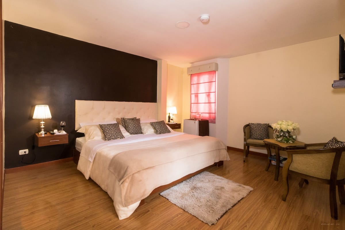 Un dormitorio con una cama grande, mesitas de noche con lámparas, una silla y una pequeña mesa con flores.