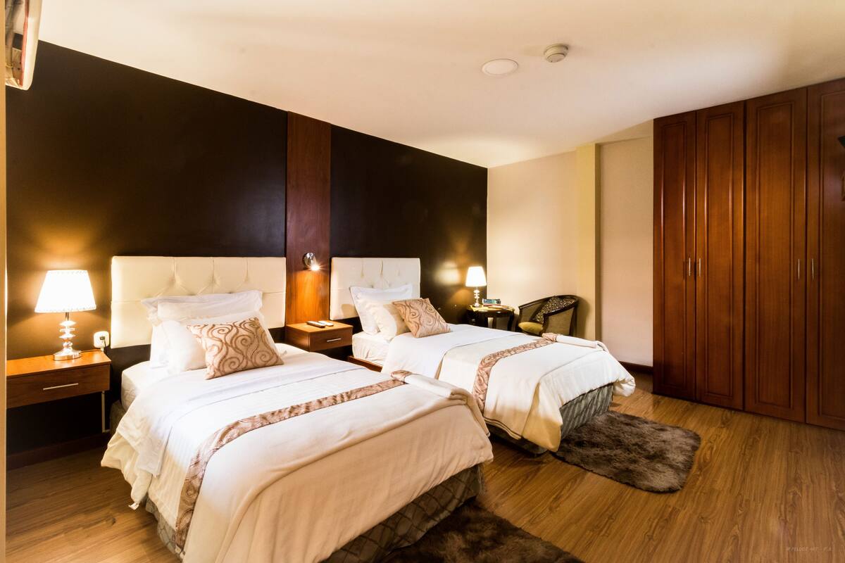 Una habitación de hotel con dos camas, un armario de madera, mesitas de noche con lámparas y una silla.