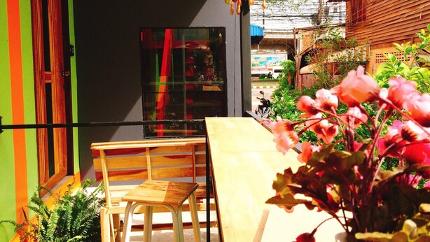 A small outdoor seating area with a wooden bench and table, surrounded by greenery and a building with a window. - Nap Corner Hostel (Phitsanulok)