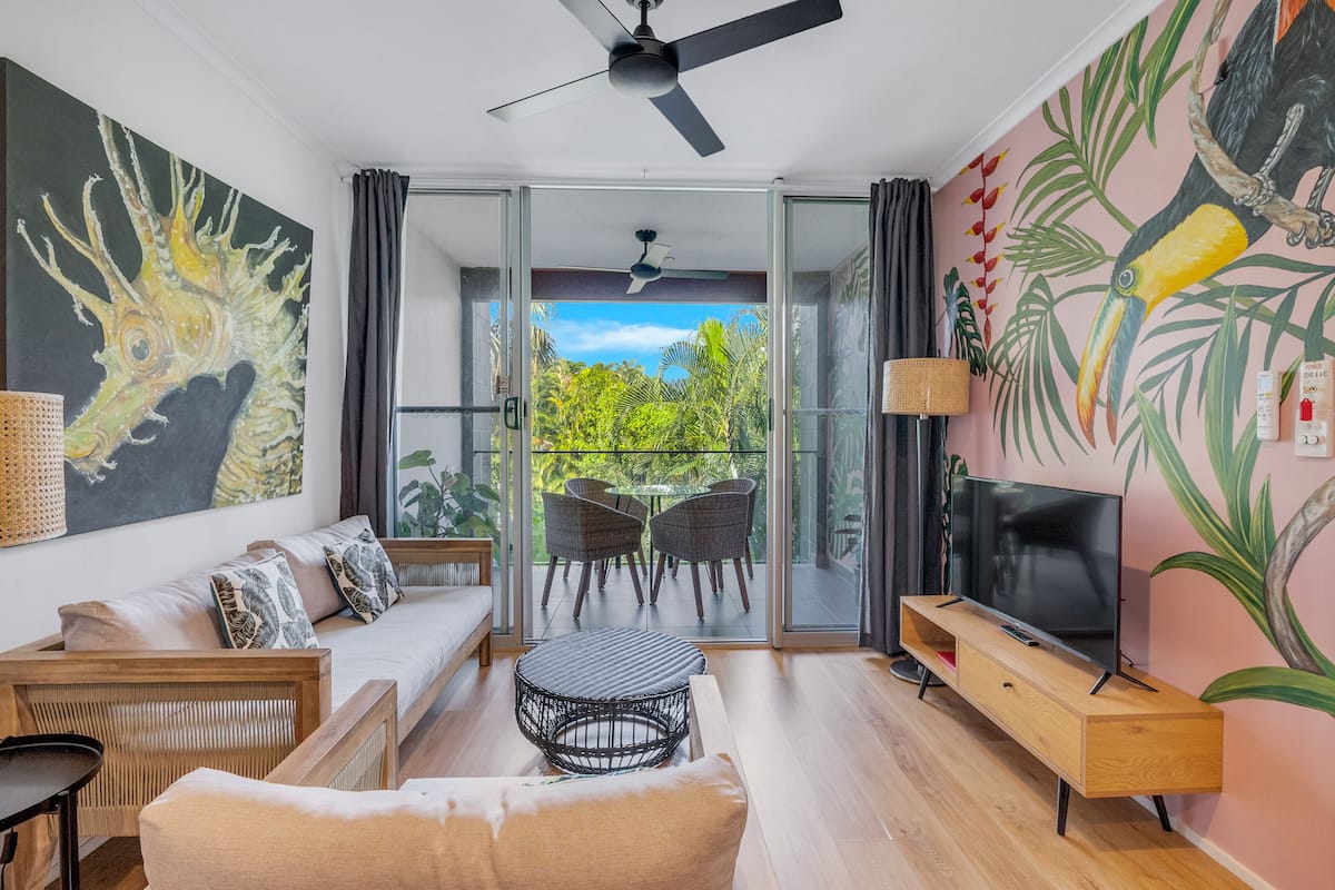 A modern living room with a sofa, TV, and a balcony with a view of greenery.