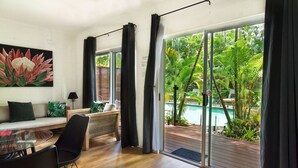 A modern living room with a glass door leading to a pool area, a sofa with cushions, a glass table, and a black chair. - The Palms at Palm Cove (Palm Cove)