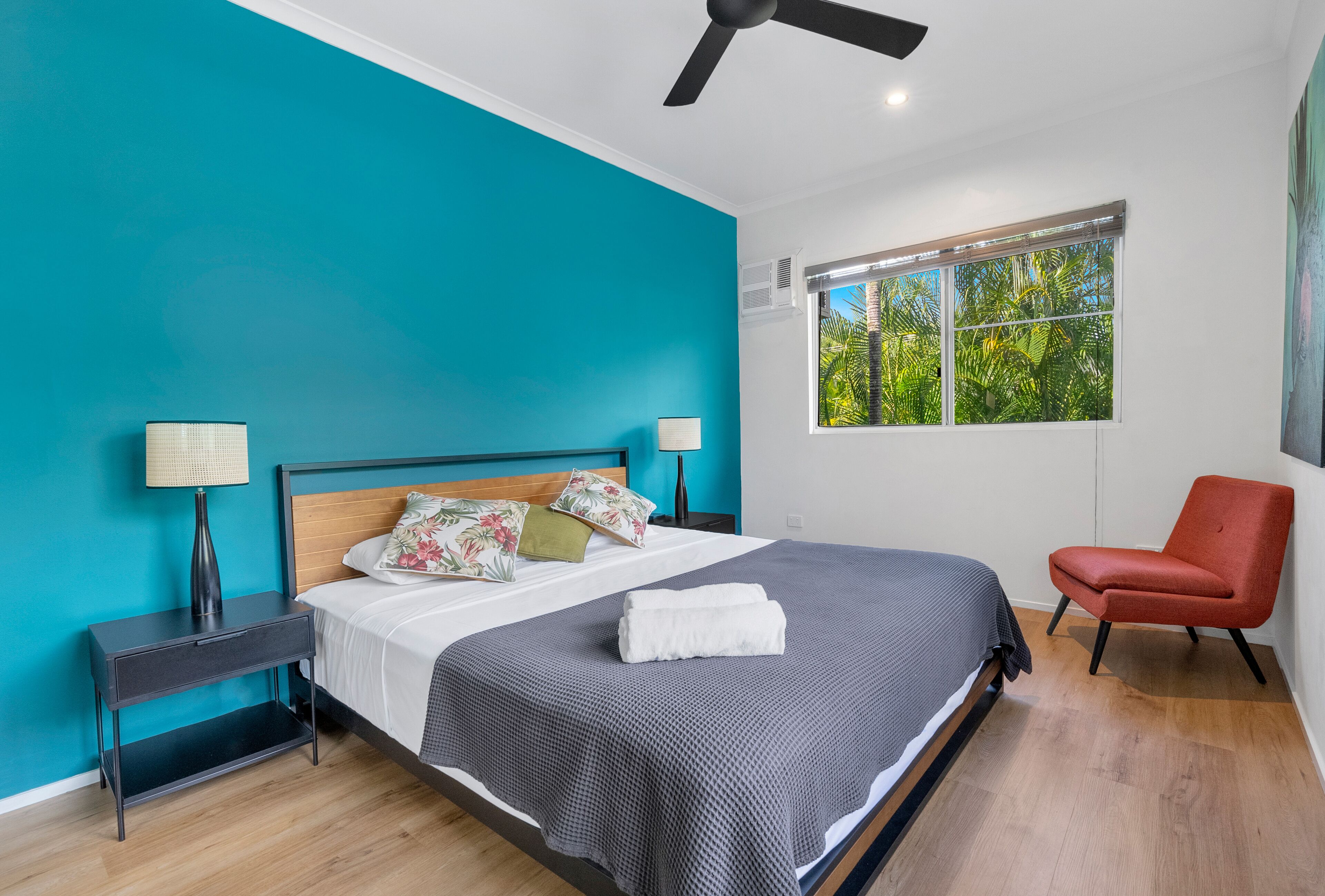 Un dormitorio moderno con una pared verde azulada, una cama con colcha gris, dos lámparas de noche, una silla roja y una ventana con vistas a la vegetación.